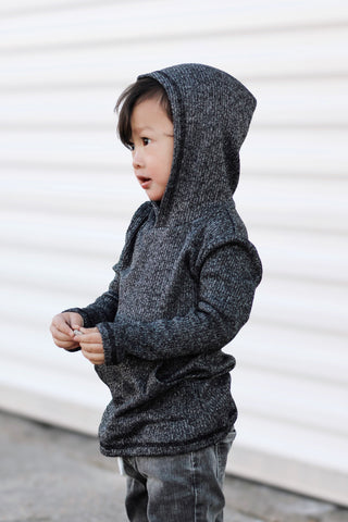 Child wearing a dark gray hooded jacket standing against a white wall.